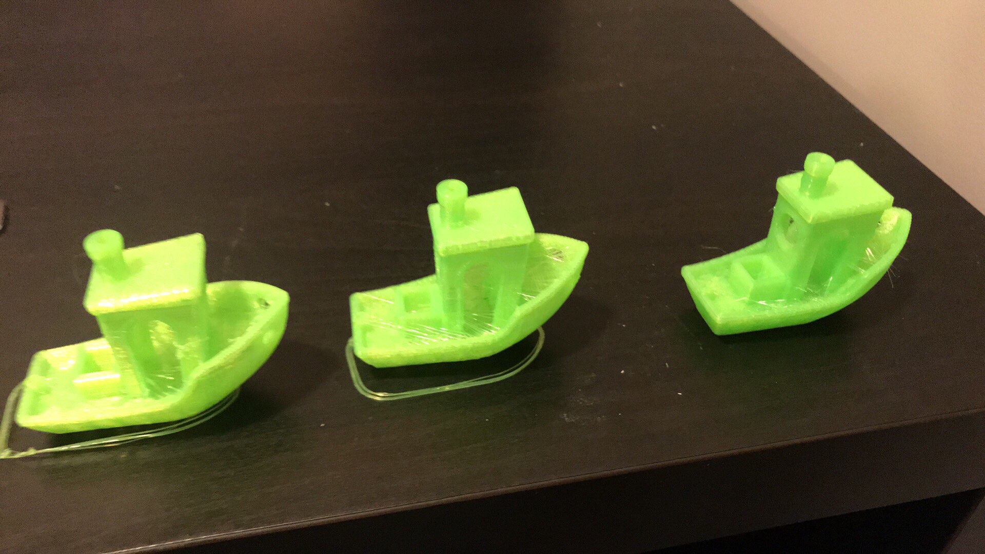 Tre grønne benchy-båter på rekke på et mørkt skrivebord. Hver båt viser samme modell printet med litt forskjellige innstillinger, med ulike mengder strenger og overflateruheter i skroget og taket.