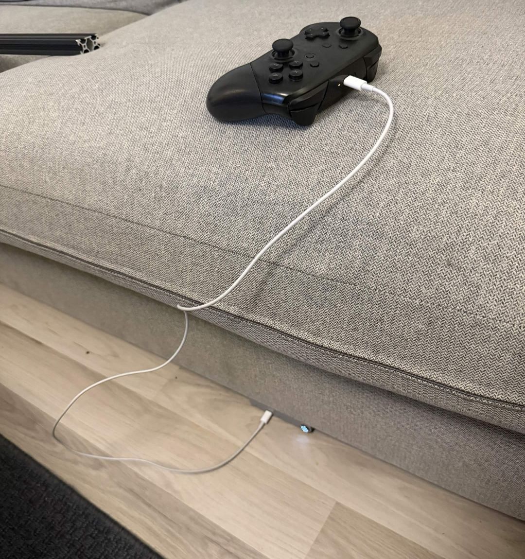 En Nintendo Switch Pro-kontroller som ligger på en sofapute og lader via en hvit kabel som går ned til stasjonen bygget inn i sofaen under.