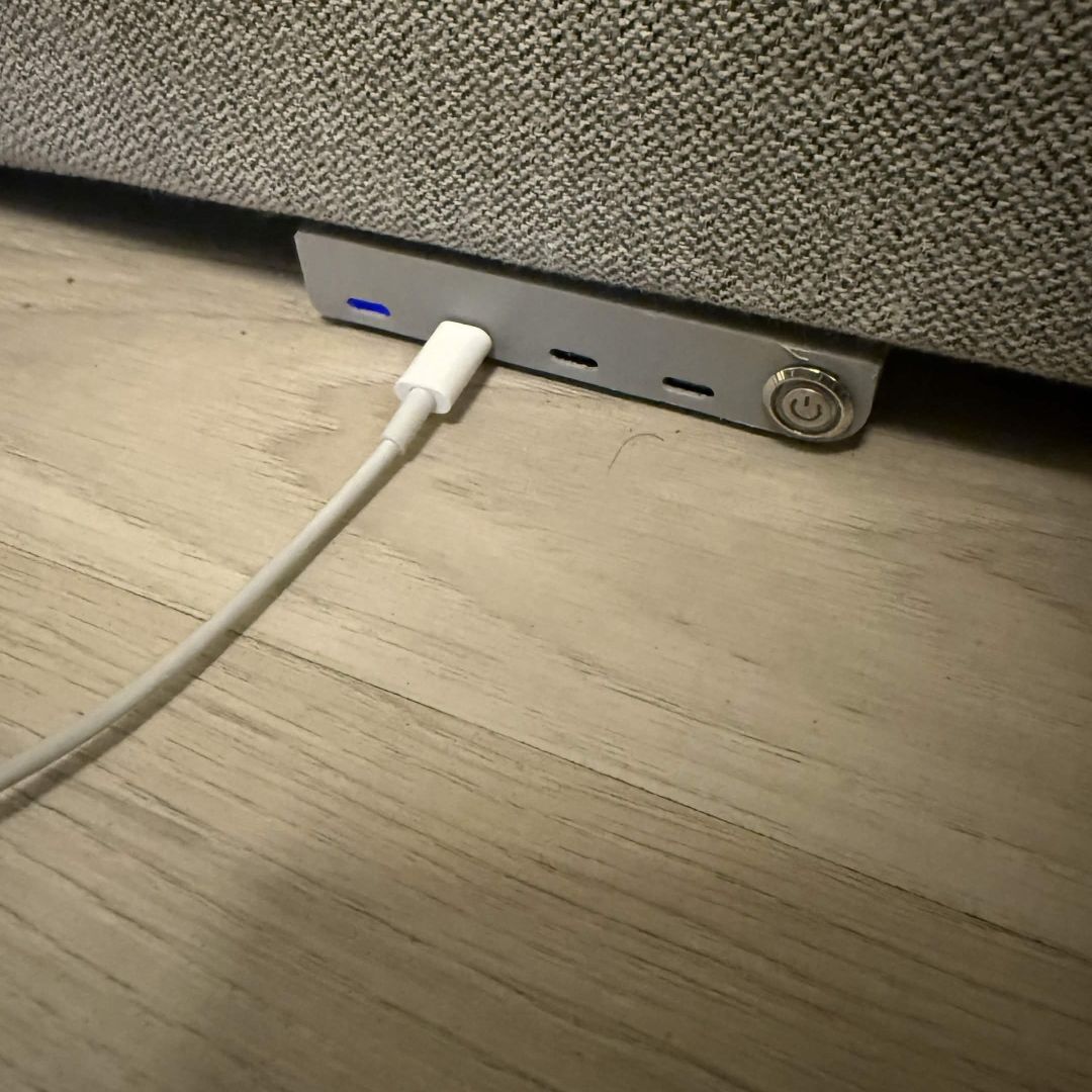 Ladestasjonen bygget inn i kanten av en grå sofa, bryteren av, en ladekabel koblet i én port.